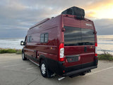 Red Winnebago Solis rv rental camper van by the ocean. Rear view showing side windows, rooftop AC, and marine backdrop.