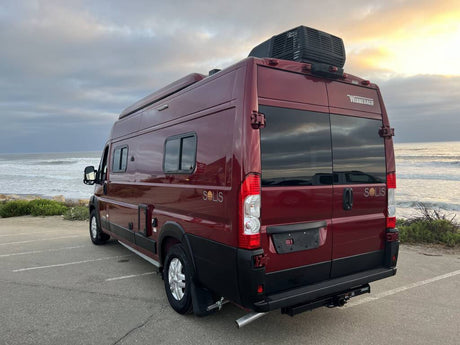 Red Winnebago Solis rv rental camper van by the ocean. Rear view showing side windows, rooftop AC, and marine backdrop.