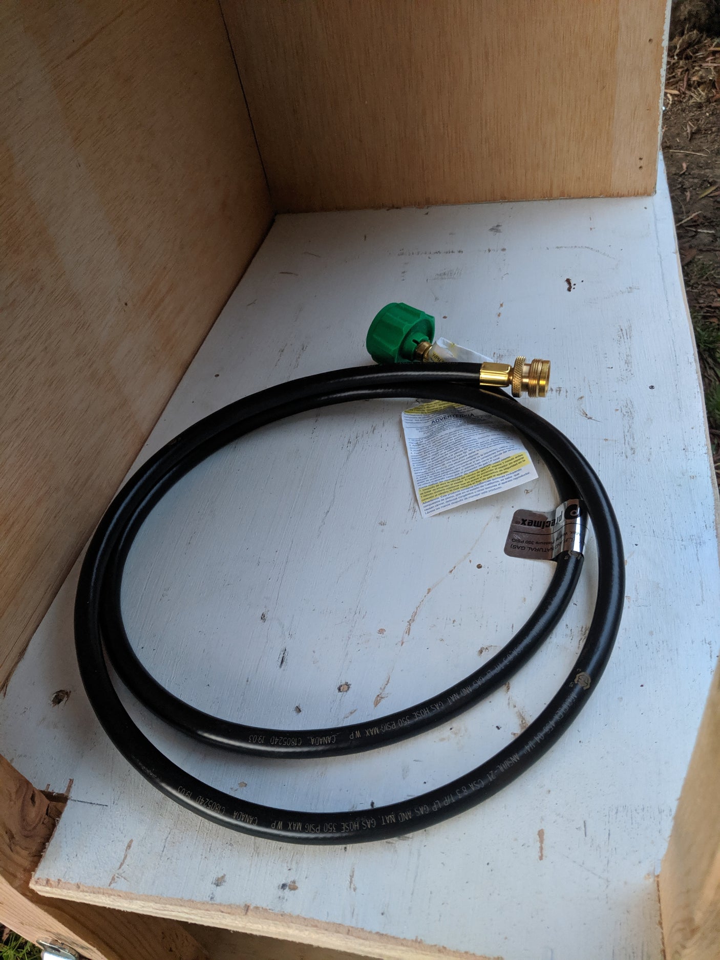 New black propane gas hose with brass fittings and green cap for an RV rental, coiled inside a wood compartment.