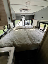 Relaxing RV rental bedroom. Features a comfortable bed with quilted blanket, overhead storage, AC, and rear windows.