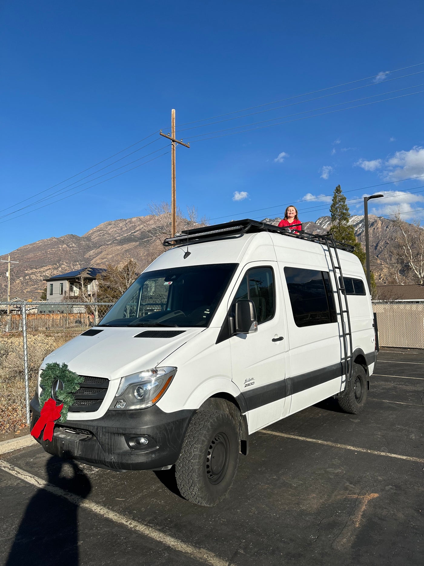 This white Sprinter RV rental features a child on its roof, a festive wreath, and a mountain backdrop. — photo 5