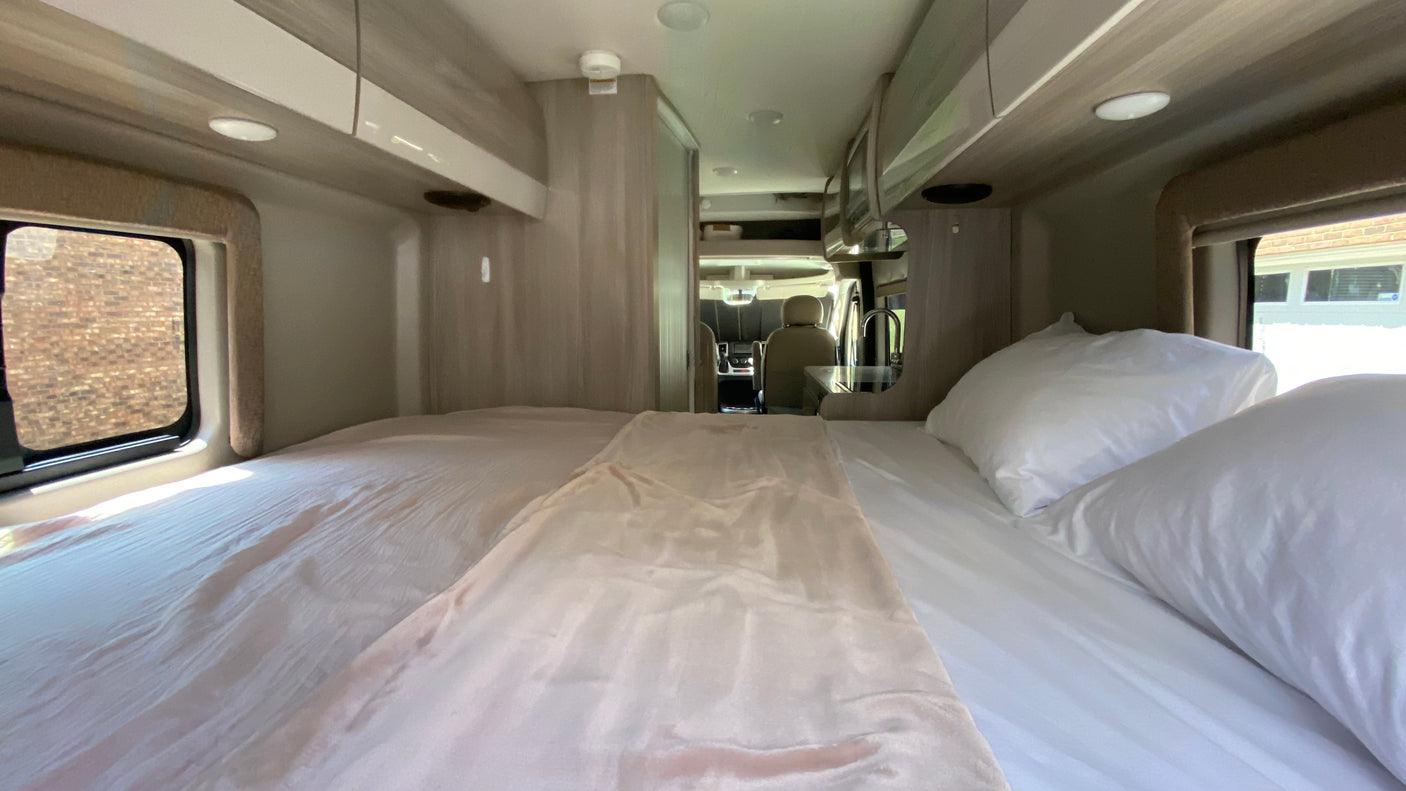View inside an RV rental: a neat bedroom with white bedding, looking towards the front driving cabin.