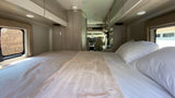 View inside an RV rental: a neat bedroom with white bedding, looking towards the front driving cabin.
