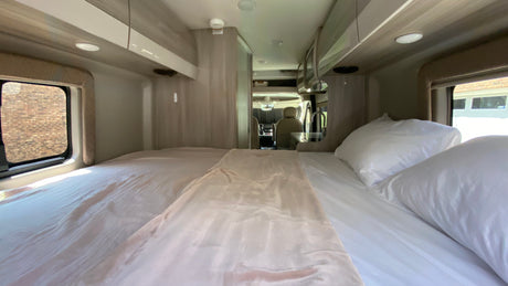 View inside an RV rental: a neat bedroom with white bedding, looking towards the front driving cabin.