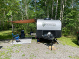 Viking Max RV rental: A travel trailer with an orange awning, chairs, and table, set up for camping in a wooded site.