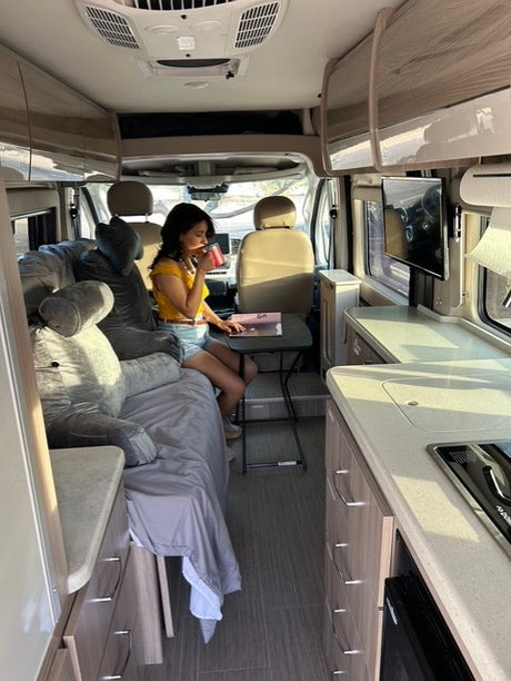 Cozy RV rental interior: woman sips a drink in a camper van, featuring a bed, kitchen, and dining spot for travel.