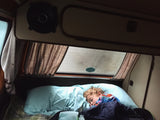A child peacefully sleeps in a cozy RV rental bed, nestled inside a camper van with curtains & speaker visible, perfect for travel.