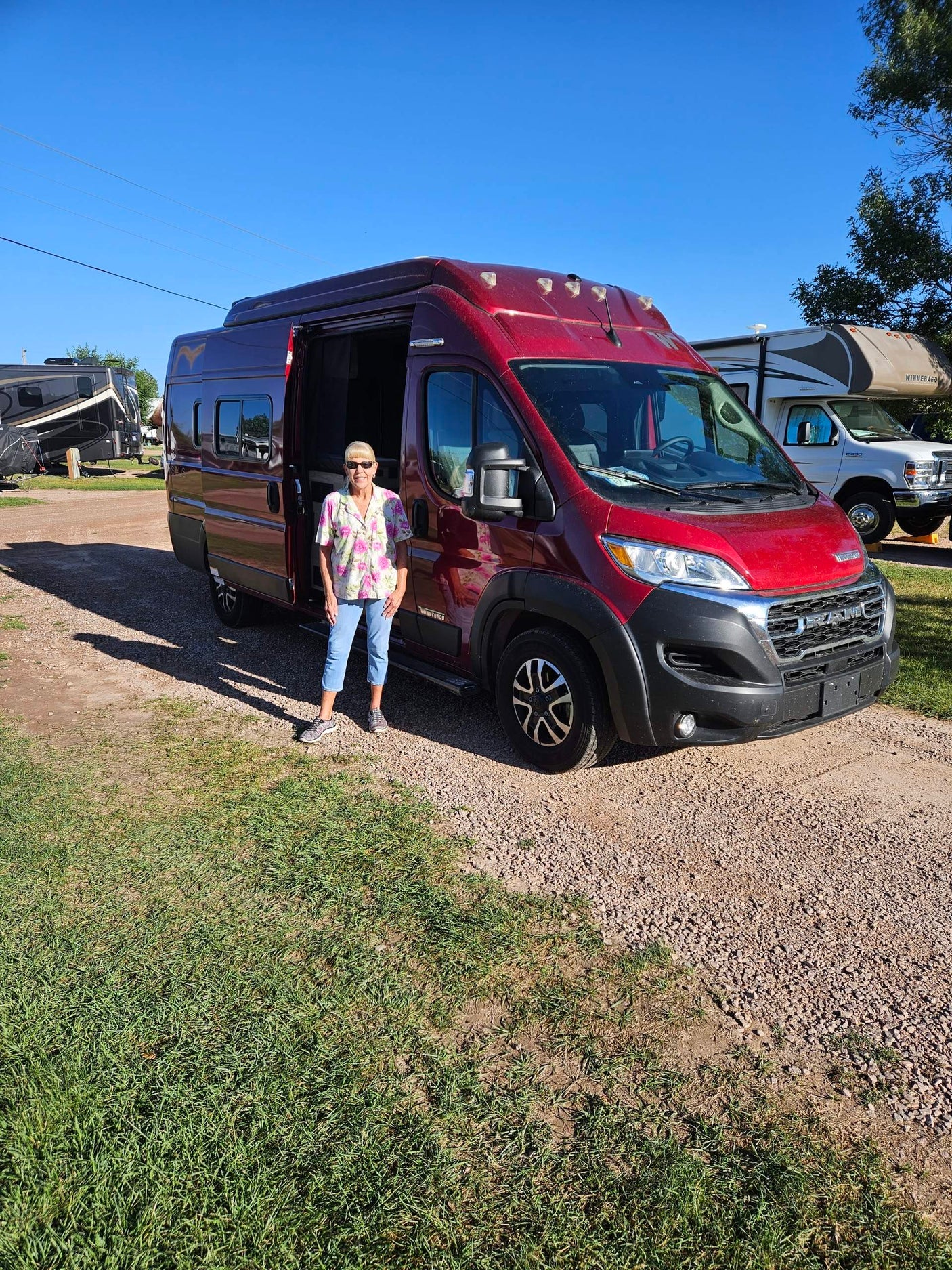 Woman with a red RV rental on a sunny day. Gravel and grass lot with other RVs in the background.