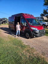 Woman with a red RV rental on a sunny day. Gravel and grass lot with other RVs in the background.