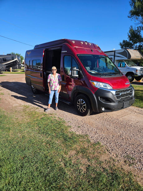 Woman with a red RV rental on a sunny day. Gravel and grass lot with other RVs in the background.
