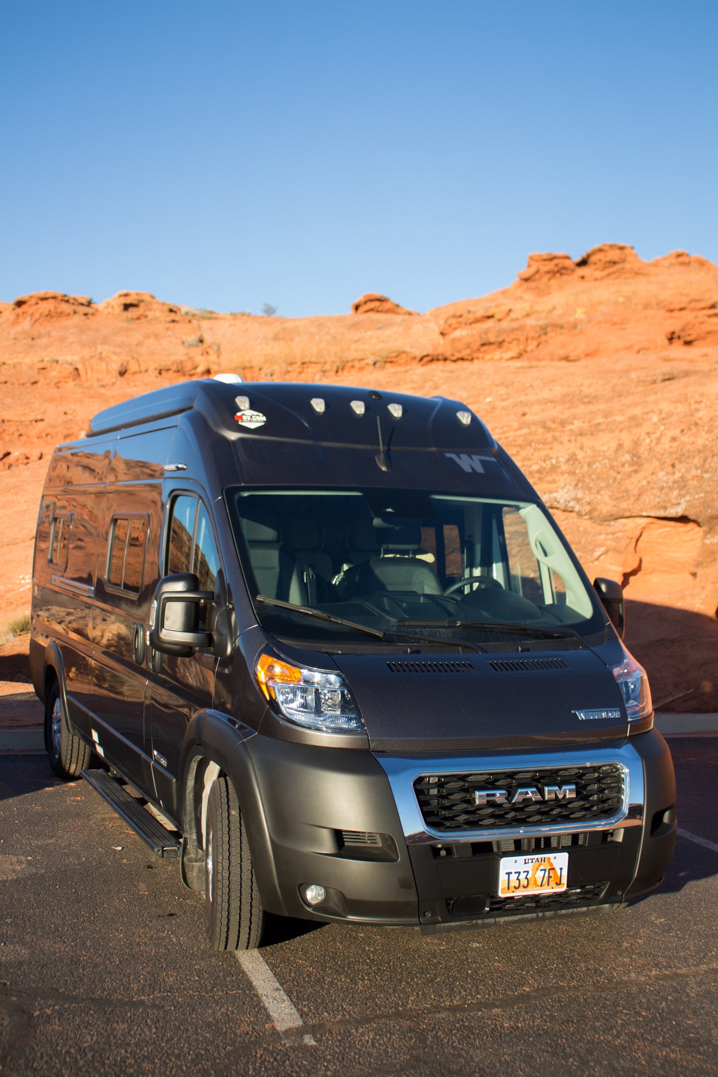 Dark grey Winnebago Ram ProMaster RV rental, parked on asphalt with red rock landscapes and a clear blue sky.