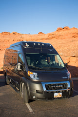 Dark grey Winnebago Ram ProMaster RV rental, parked on asphalt with red rock landscapes and a clear blue sky.
