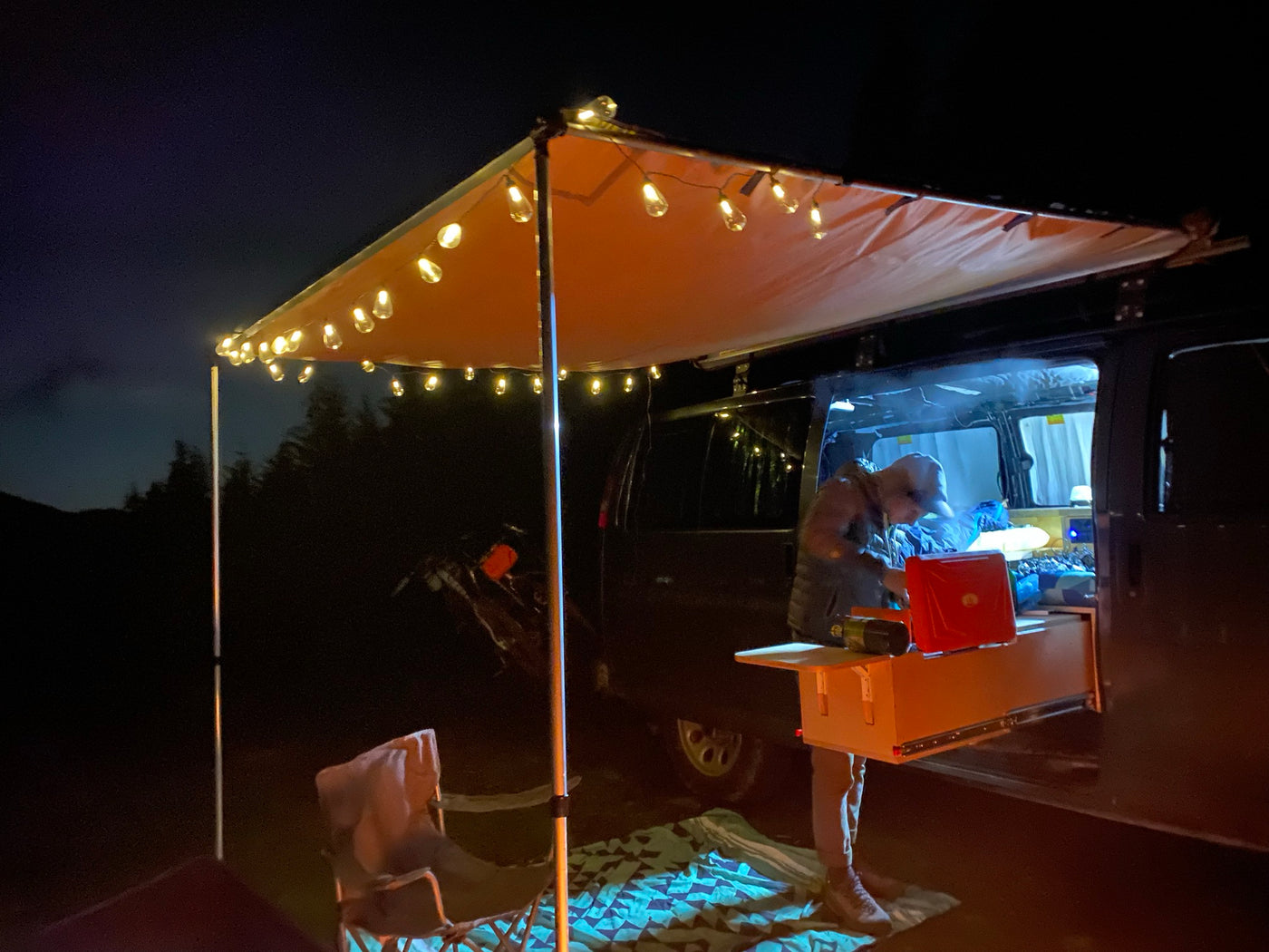 Nighttime RV rental setup: A person prepares food from a pull-out kitchen under a brightly lit awning with string lights. — photo 9