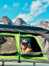 Smiling woman in an RV rental rooftop tent enjoys majestic mountain views on a sunny day.