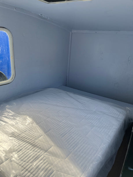 Inside an RV rental: a pristine mattress covered in clear plastic, set within a compact, light grey sleeping area with a small window.