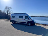 A silver Winnebago Solis RV rental is parked on an asphalt road next to a calm lake, with trees in the background under a sunny sky.