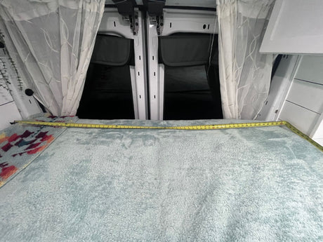 Cozy RV rental sleeping area: Bed with measuring tape, sheer curtains frame rear van doors.