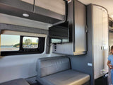 Cozy RV rental interior with bench seating, TV, overhead cabinets, and a window view of the beach.
