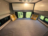 Interior of an **RV rental** showing a neatly made bed with plaid pillows, overhead cabinets, and bright windows with outdoor views.