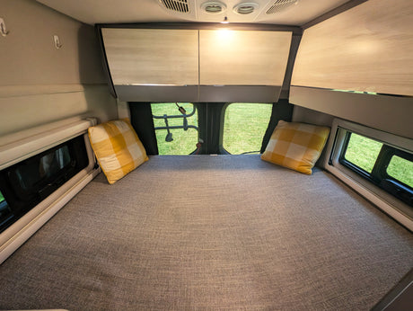 Interior of an **RV rental** showing a neatly made bed with plaid pillows, overhead cabinets, and bright windows with outdoor views.
