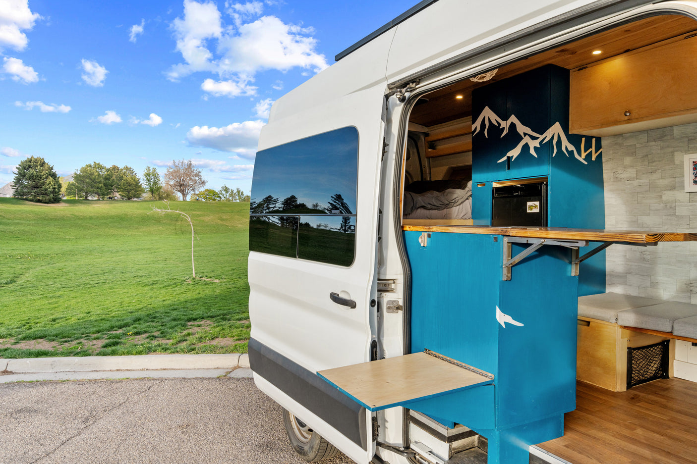 Modern RV rental campervan interior with blue & wood accents, fold-out tables, and a serene outdoor view.