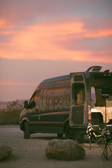 This 'the tripsit' RV rental camper van, parked at sunset, has its rear doors open, revealing a cozy interior in a desert setting.