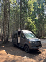 Grey camper van, side door open, parked on a dirt road in a sunny forest. Great for an RV rental adventure!