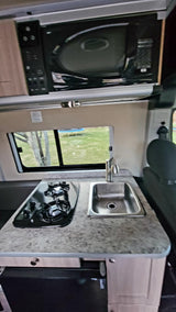 Compact RV rental kitchenette: microwave, two-burner stove, and sink. Window view of outdoors.