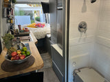 Bright RV rental interior features a compact kitchen with fruit, a comfy bed, and a full shower/toilet for comfortable mobile living.