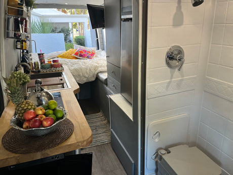 Bright RV rental interior features a compact kitchen with fruit, a comfy bed, and a full shower/toilet for comfortable mobile living.