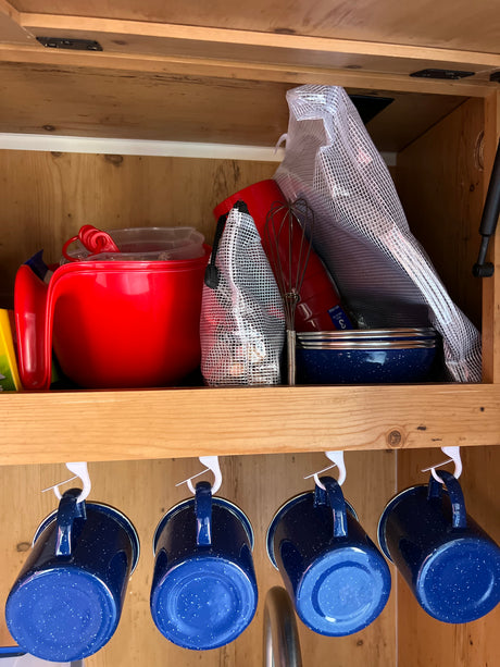 An RV rental's organized kitchen cabinet showing red bowls, blue enamel mugs hanging, and cooking essentials, perfect for travel.