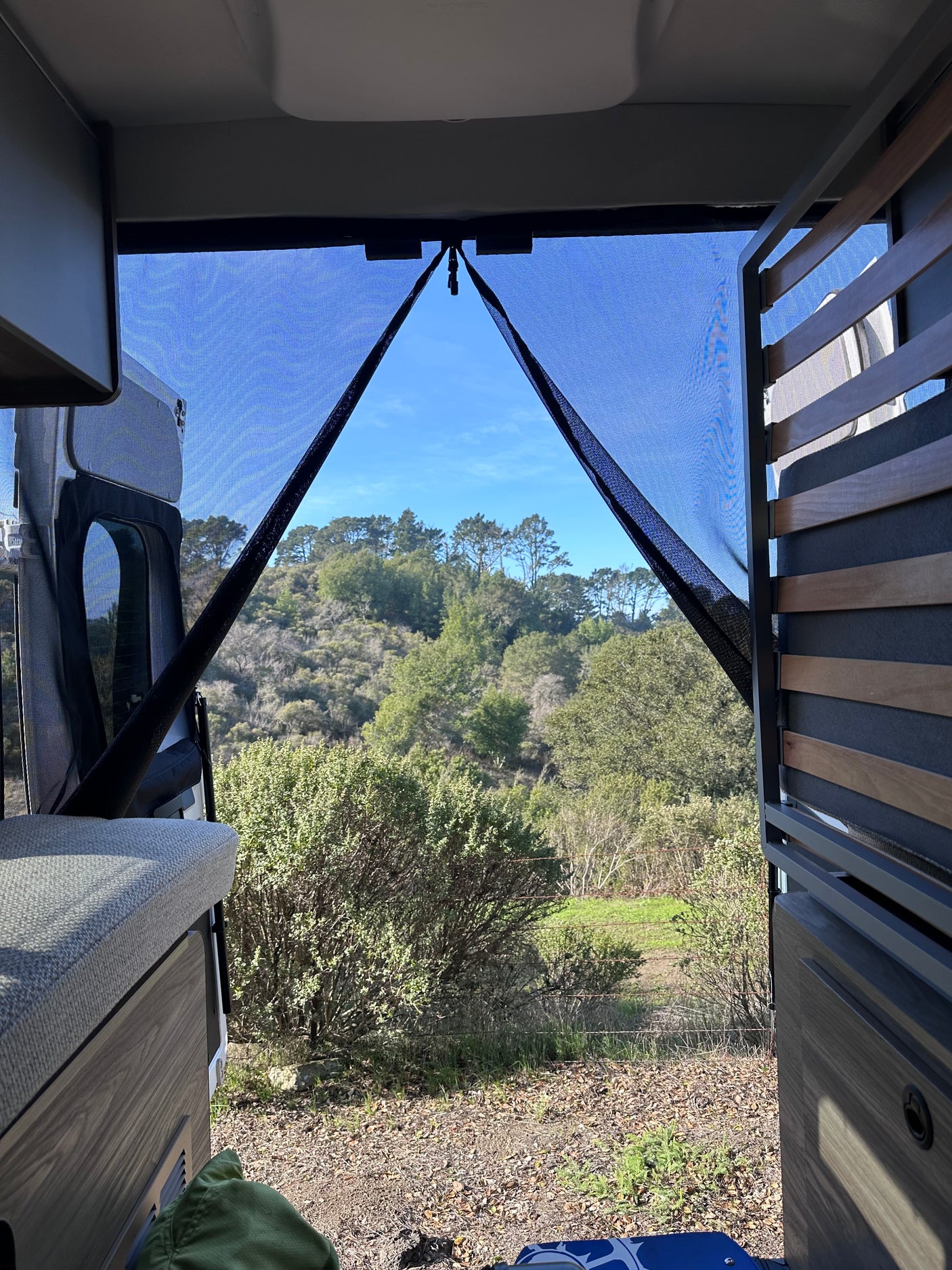 Peering through an RV rental's screen door at a serene natural landscape with lush green hills and trees. Van camping bliss!