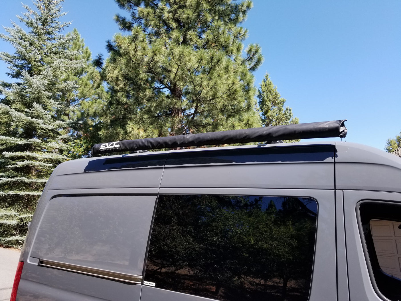 Grey RV rental van with a black CVC roof awning, parked outdoors under clear skies.