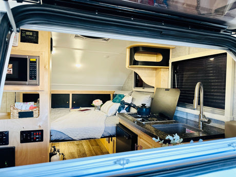Step inside this modern RV rental: a cozy bed, compact kitchen with sink/stove, and control panels for comfort on the go.