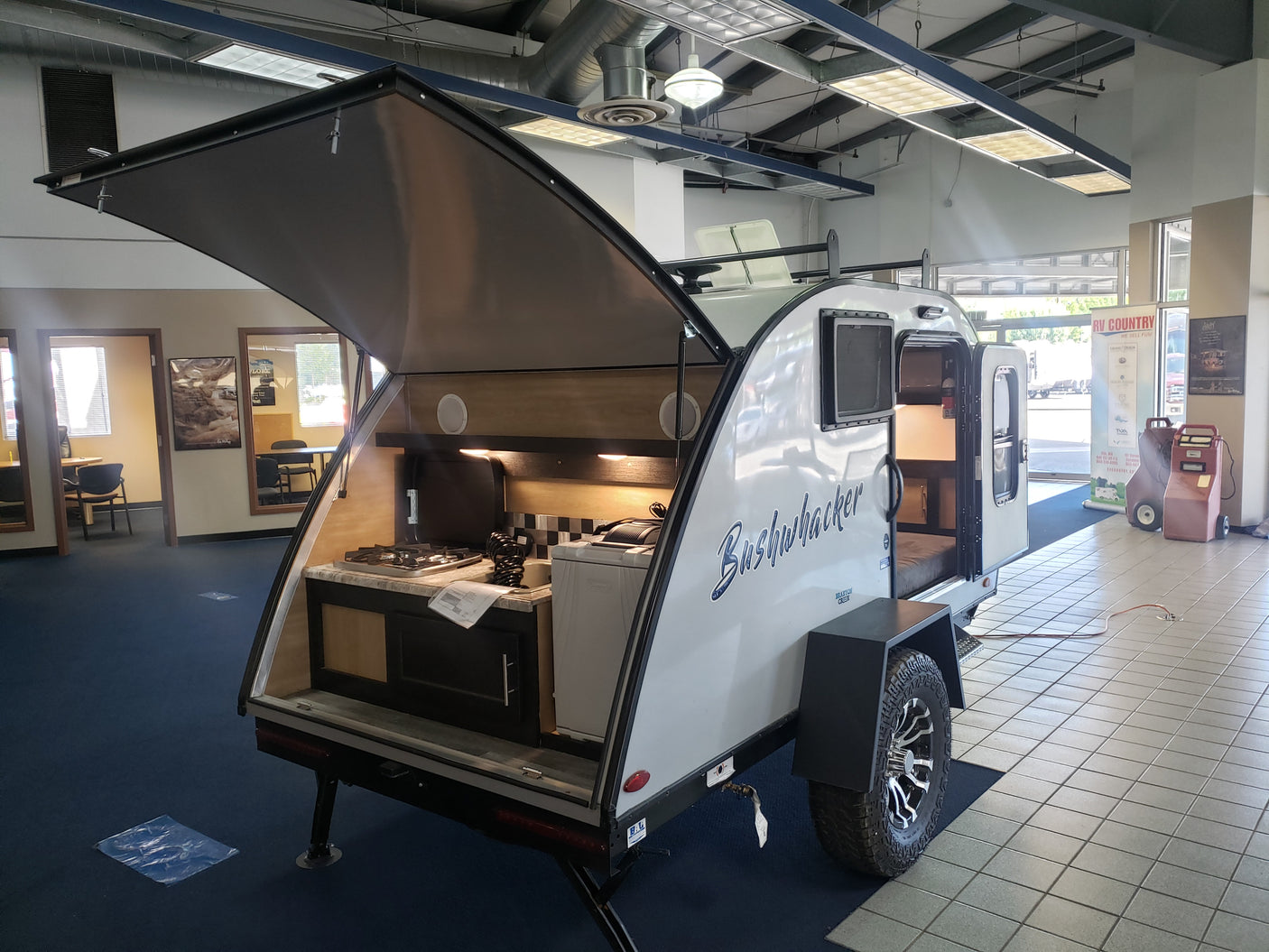 Bushwhacker teardrop trailer (compact RV rental option) with open rear kitchen and side entrance, displayed in a showroom.
