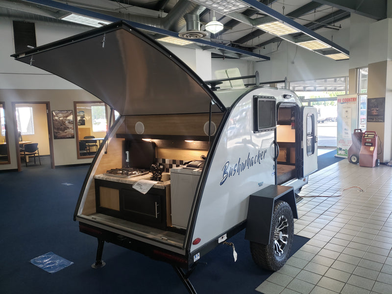 Bushwhacker teardrop trailer (compact RV rental option) with open rear kitchen and side entrance, displayed in a showroom. — photo 3