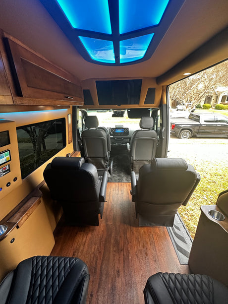 Spacious RV rental interior: view from the rear seats towards the front, with custom blue ceiling lights, wood floors, and captain's chairs.