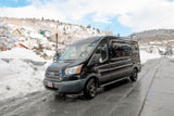 Black Ford Transit van for winter RV rental trips, parked on a snow-covered street in a picturesque mountain town.