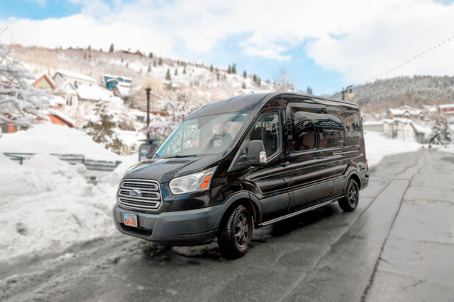Black Ford Transit van for winter RV rental trips, parked on a snow-covered street in a picturesque mountain town.