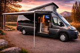 Experience an RV rental adventure! This gray camper van, with an extended awning and illuminated interior TV, sits by a scenic lake at sunset.