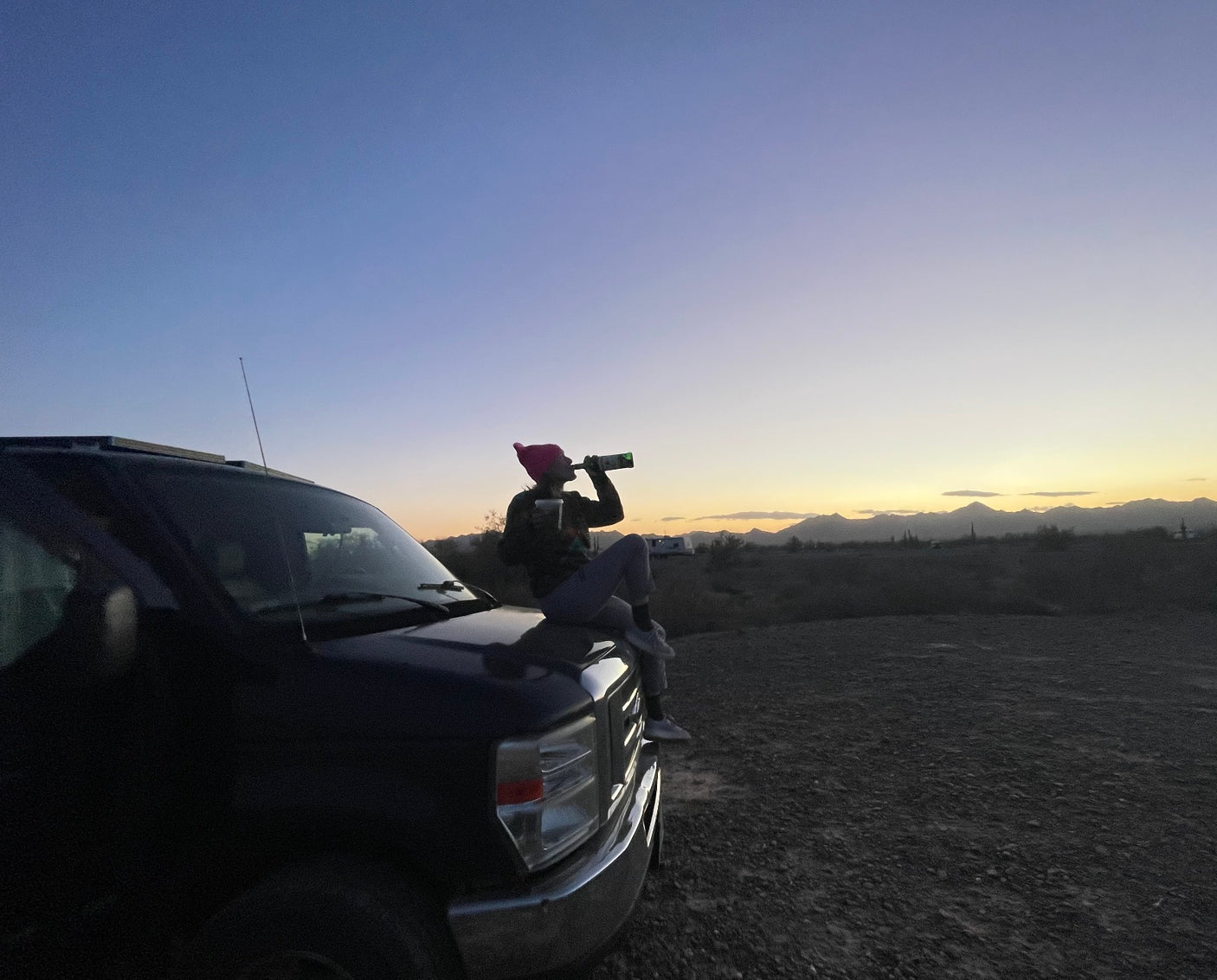 Relaxing on an RV rental van at desert sunset. Person drinks from bottle; mountains in view.