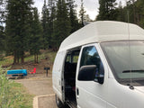 RV rental camper van parked at a scenic campsite with picnic table and grill among tall trees.