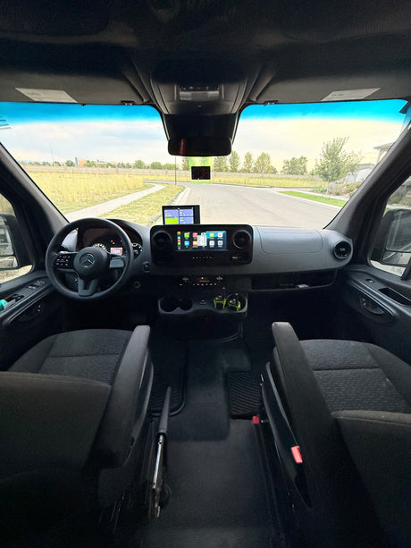 RV rental cockpit view: Mercedes dashboard, dual screens (Apple CarPlay), steering wheel, and radios. Outside, a road and fields.