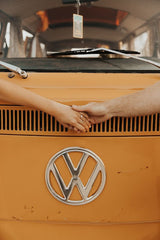 A couple's hands connect over the grille of a vibrant orange vintage VW camper van, ideal for a memorable RV rental adventure.
