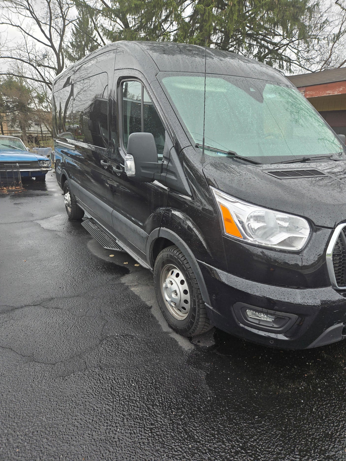 Black high-roof van, suitable for RV rental, parked on wet pavement with side steps and rain droplets.