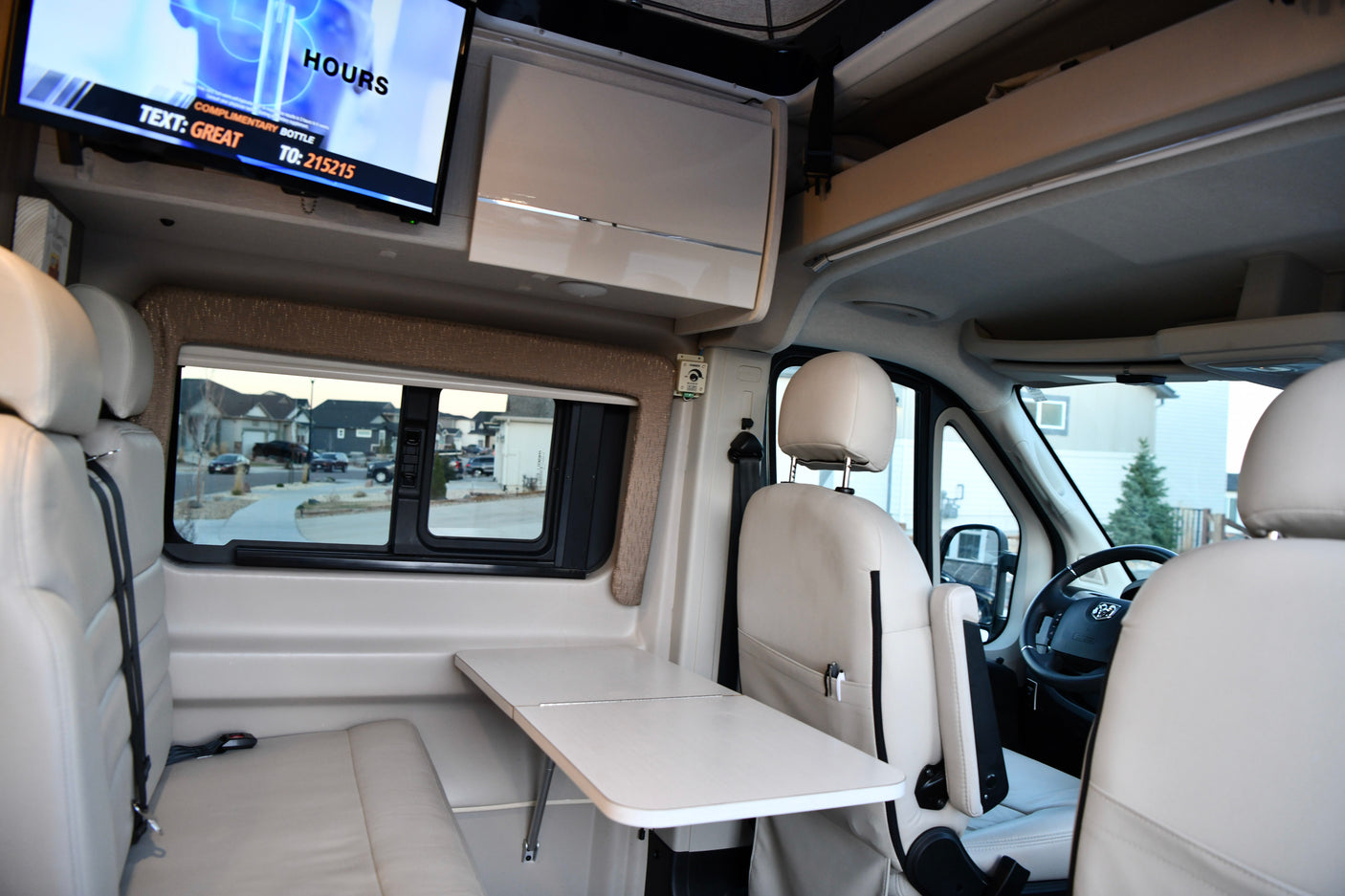 Bright rv rental interior: light seating, adjustable table, and TV. A residential street view is visible outside the window.
