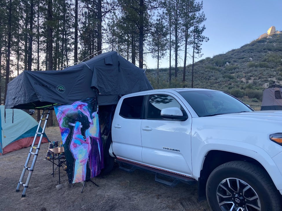 White truck with Nomadic rooftop tent set up for camping in the woods. A versatile choice for outdoor stays, similar to what you'd find in an rv rental.