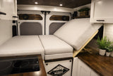 Modern RV rental camper van interior: convertible bed, cooktop, and custom wood details. Speakers visible in rear doors.
