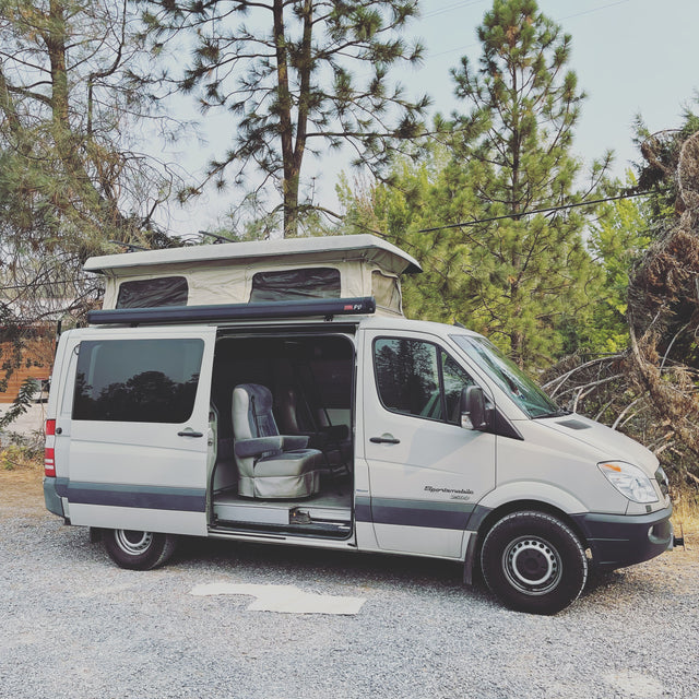Silver Sprinter camper van with pop-top roof raised and side door open, parked on gravel. Great for RV rental exploration.