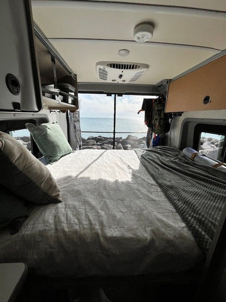 Cozy RV rental interior: a comfy bed facing rear windows with a stunning ocean and rocky shore view.